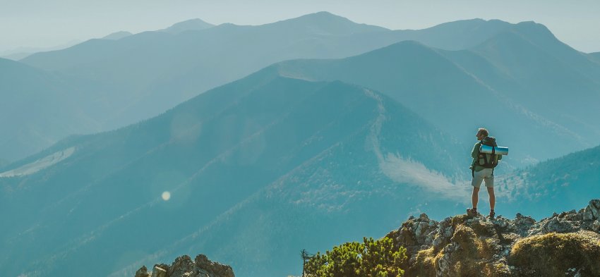 Персона с рюкзаком на фоне горной цепи, занимающаяся походами и фотографией.