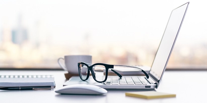 Очки на столе возле ноутбука, компьютерной мыши, чашки и другой электроники.