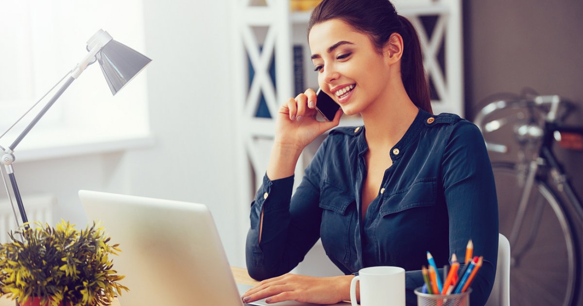 Женщина радуется, держа ноутбук, сидя у растения с чашкой кофе, взгляд на камеру.