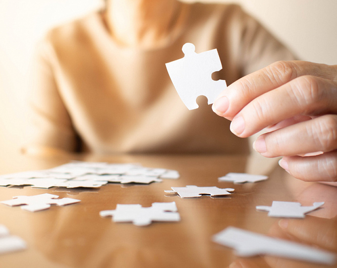 Человек собирает пазл за обеденным столом, на фоне мебели и игры.