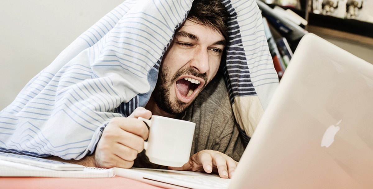 Мужчина с открытым ртом зевает, держа чашку и работая на ноутбуке под одеялом.