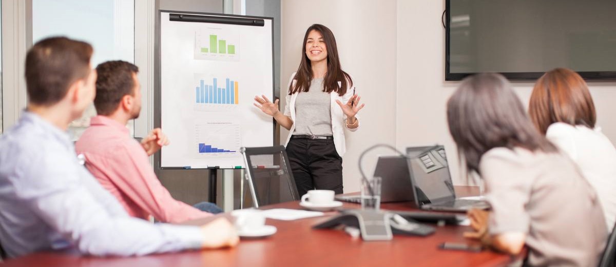 Взрослая женщина сидит за ноутбуком перед монитором и белой доской.