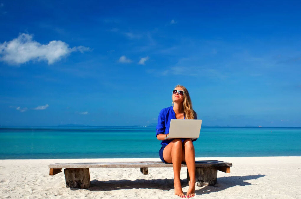 Человек сидит на пляже в летней одежде с ноутбуком и солнечными очками.