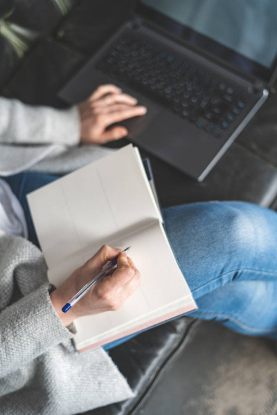 Человек пишет в книге на ноутбуке с помощью ручки и компьютера.