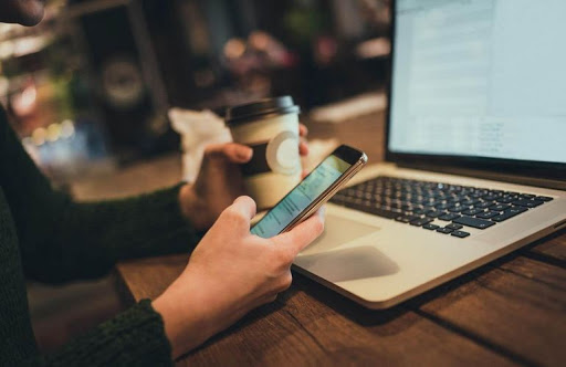 Электроника: человек пишет на ноутбуке с мобильным телефоном рядом и стаканчиком.