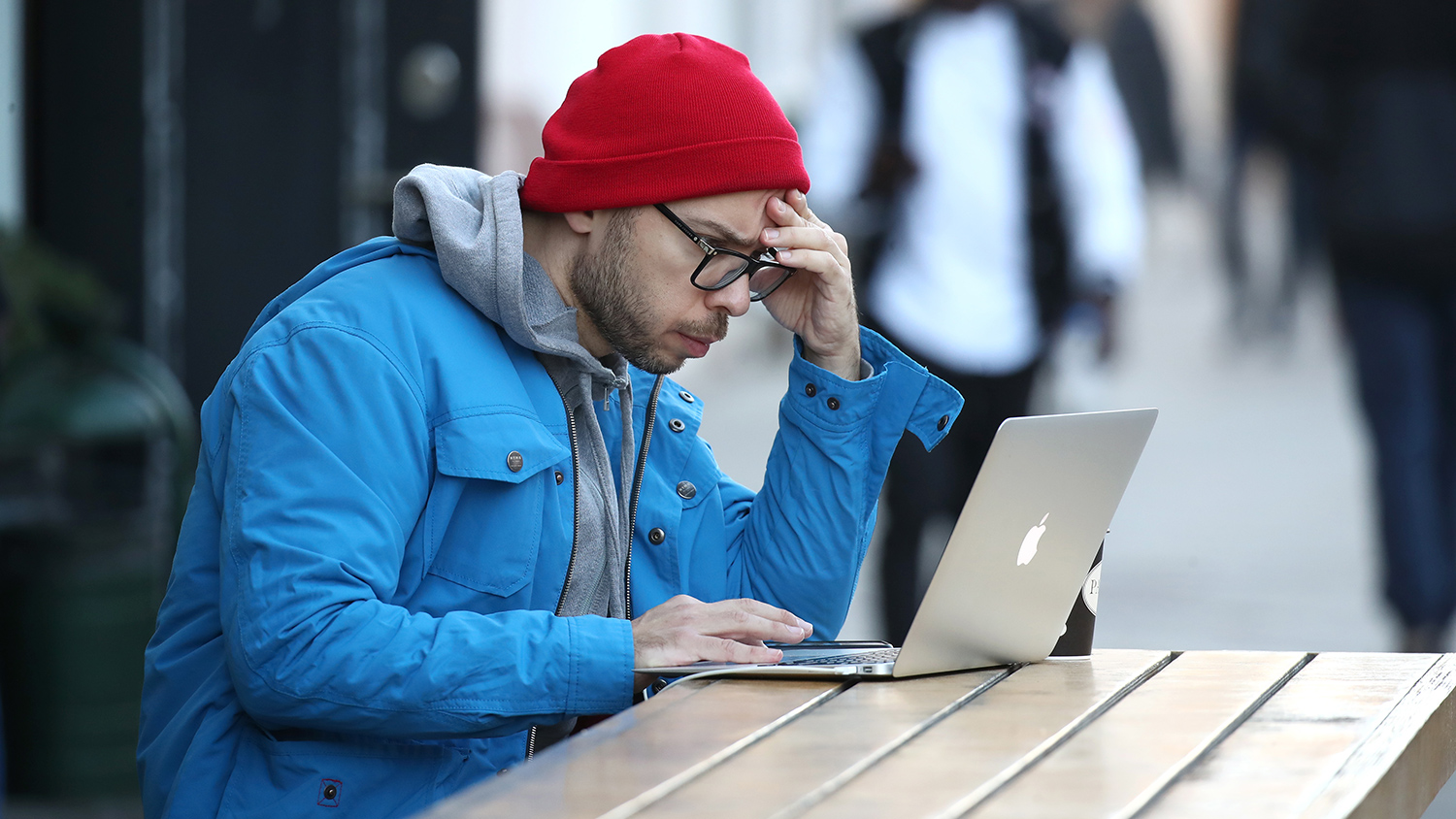 Человек мужчина в кепке совы, очках, с грустным выражением лица, работает за ноутбуком.