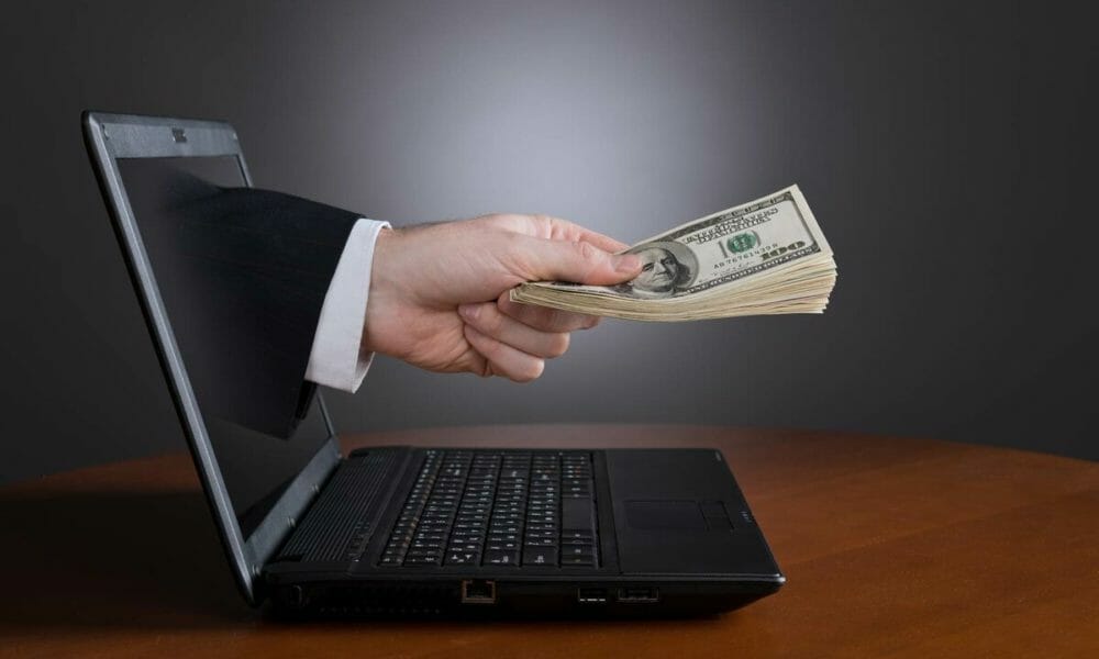 Человек сидит за ноутбуком, в фоне компьютеры, электроника, деньги.