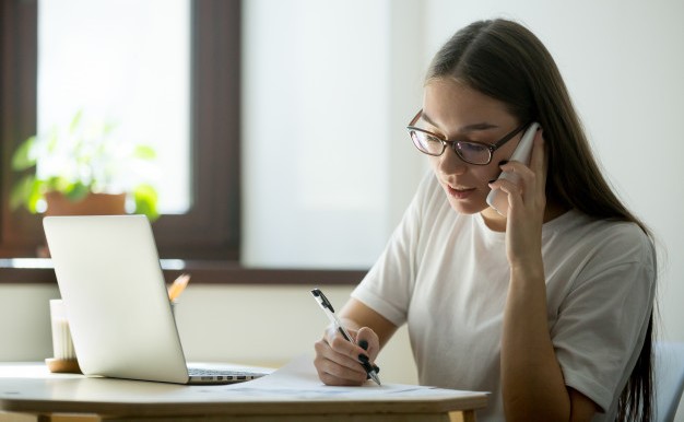 Женщина с галстуком и очками пишет на ноутбуке с телефоном.