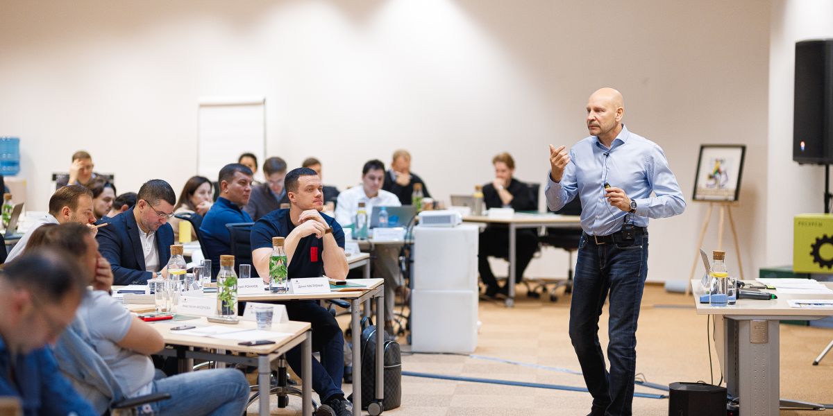 Мужчина в джинсах стоит среди толпы людей в помещении.