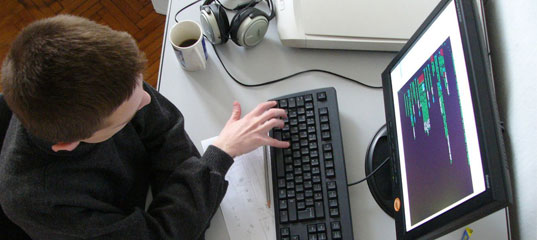 Человек за компьютерным столом с монитором, клавиатурой, наушниками и чашкой.