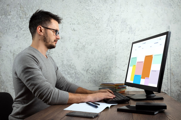 Клавиатура, монитор, мужчина за столом с мобильным телефоном, ручка и стул.