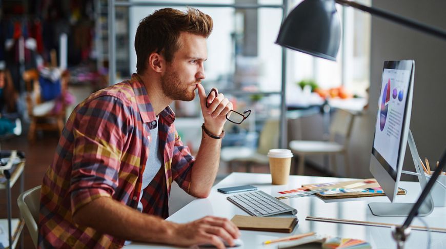 Мужчина сидит за компьютером, взгляд на монитор, рядом мобильный телефон.