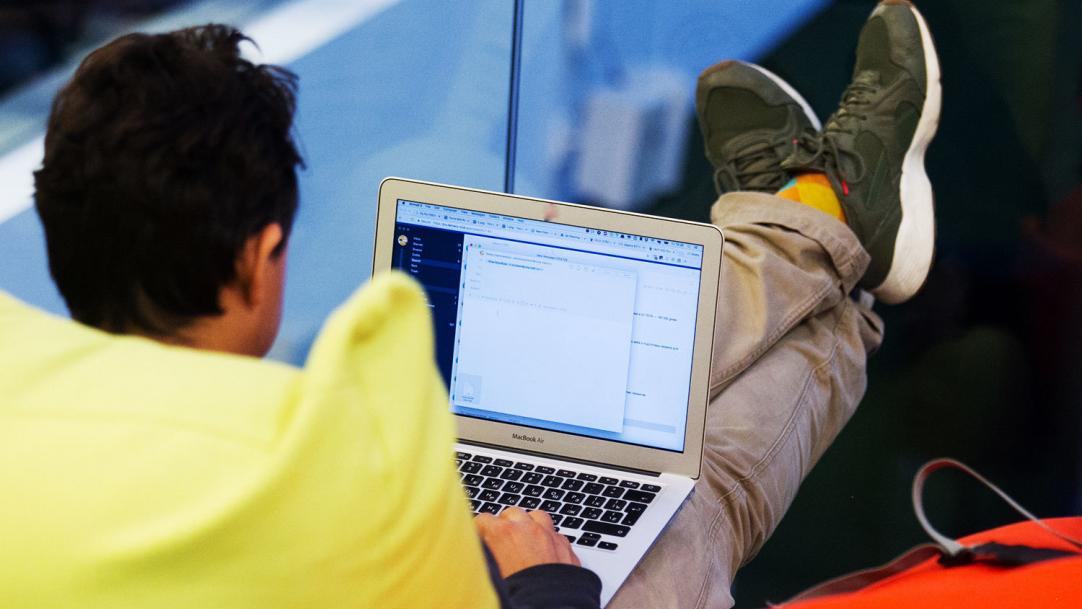 Человек в кроссовках за ноутбуком с монитором и клавиатурой на заднем плане.