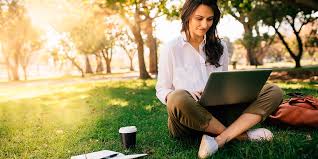 Женщина в блузке сидит на траве, читает и работает на ноутбуке.