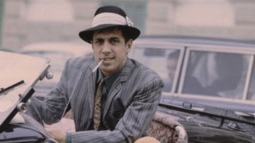 Мужчина в шляпе и пальто, стоит возле автомобиля с бейсбольной кепкой и номерным знаком.