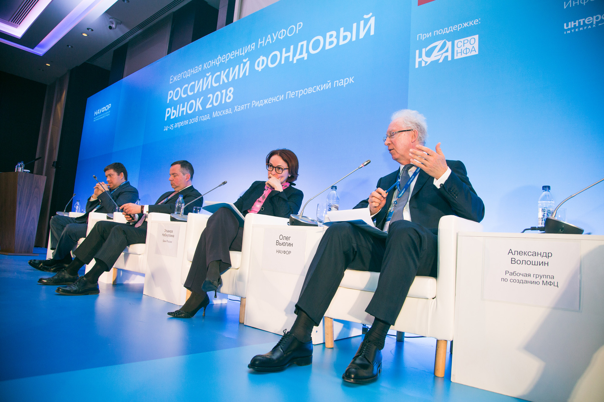 Толпа взрослых людей, мужчина в очках и туфлях среди аудитории на выступлении.