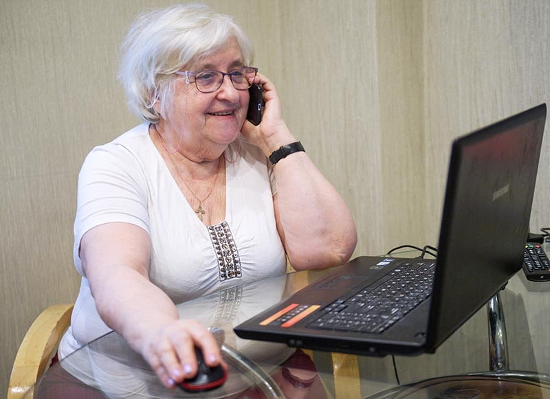 Женщина-пенсионерка смотрит на мобильный телефон на фоне ноутбука и ПК.