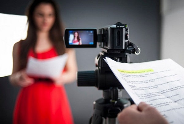 Фотографирующая взрослая женщина с видеокамерой на штативе перед монитором.