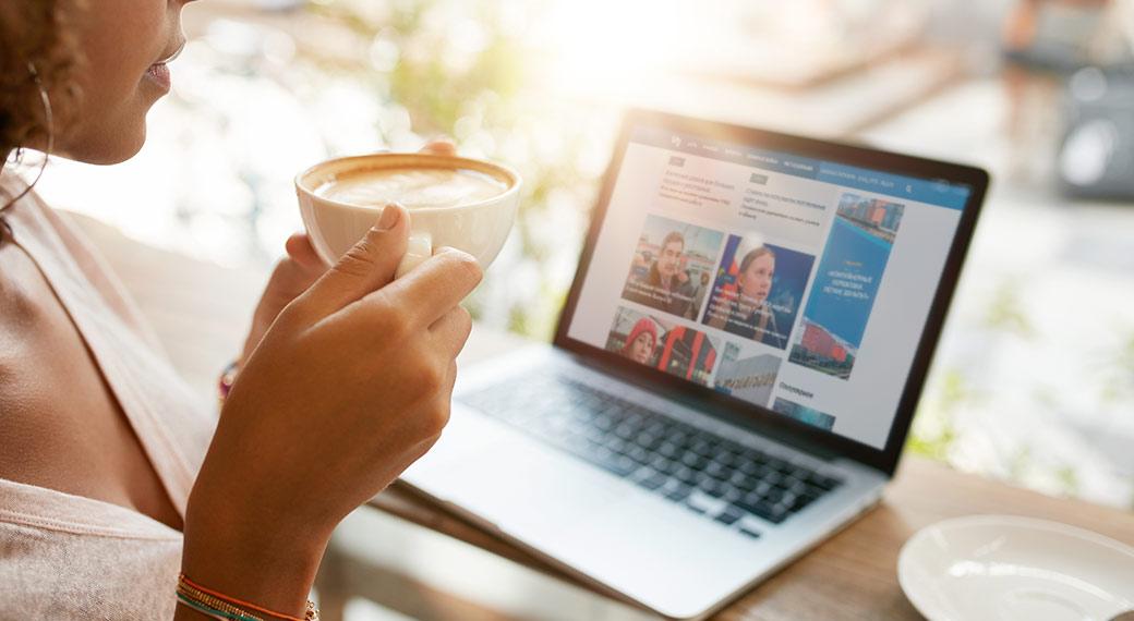 Женщина сидит за компьютером и пьет кофе, на столе лэптоп и клавиатура.