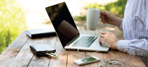 Ноутбук, мобильный телефон, стол, персона, ручка и стакан на рабочем столе.