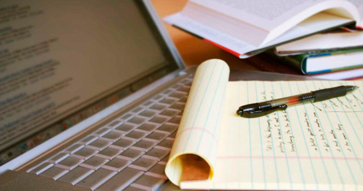 Человек пишет в книге с помощью ручки на ноутбуке с текстом на экране.