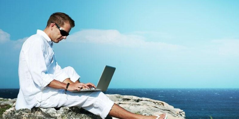 Мужчина в пляжной одежде сидит с ноутбуком на свежем воздухе.