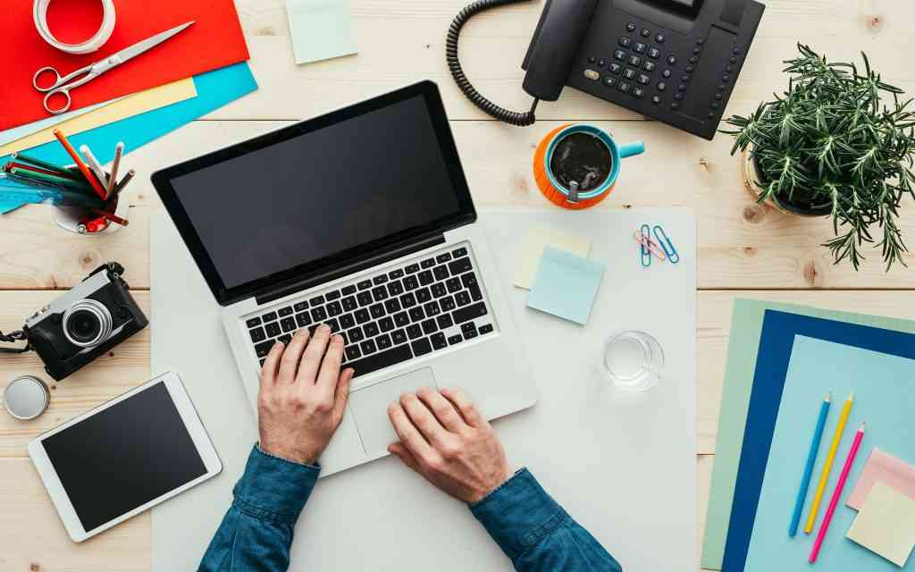 Компьютер на столе с электроникой, ноутбук, мебелью, телефоном и растением.