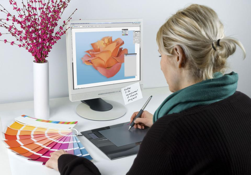 Женщина сидит у стола с компьютером, держащая ручку, смотрит на монитор.