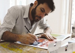 Взрослый мужчина сидит за столом рядом с живописью и мебелью.