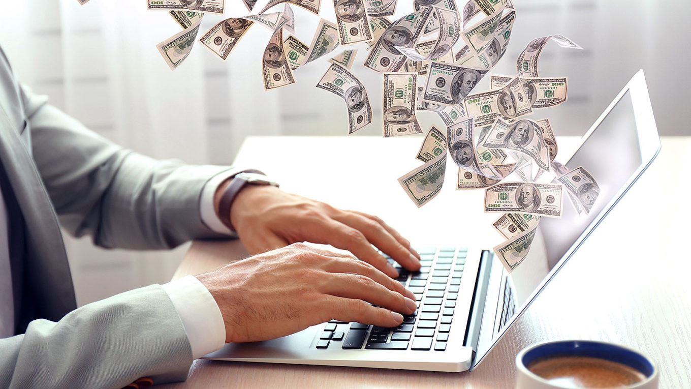 Мужчина с лэптопом, чашкой кофе и клавиатурой, держащий деньги, с видимым лицом.