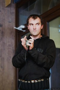 Портрет человека с микрофоном на фоне толпы, в пальто и куртке с капюшоном.