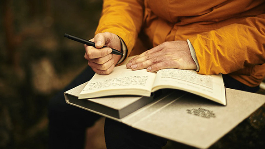 Человек мужского пола сидит за столом и пишет в книгу ручкой.