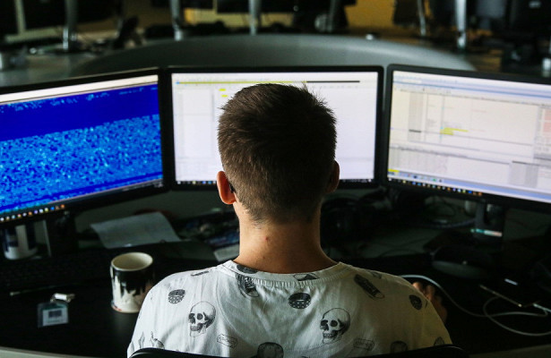 Человек работает на компьютере за столом с монитором, электроникой и чашкой кофе.