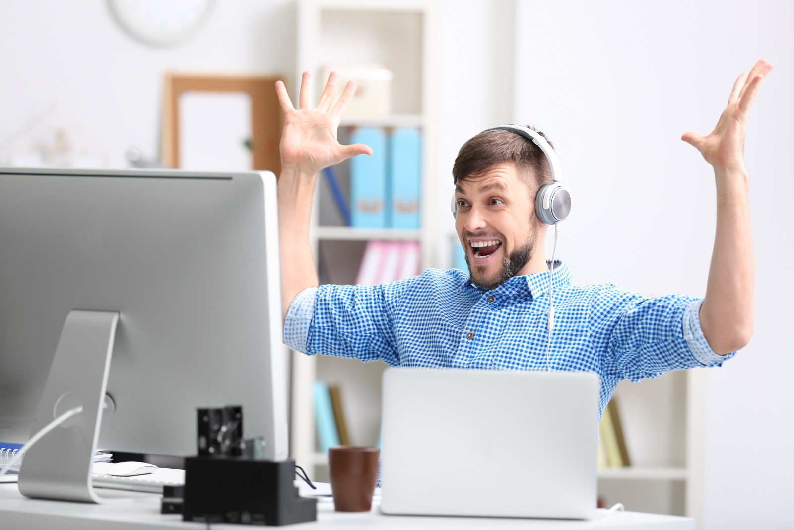 Мужчина радостно улыбается, смотря на ноутбук, в наушниках, перед монитором.