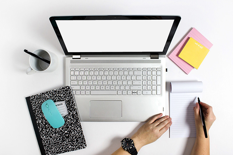 Человек работает на ноутбуке за столом с клавиатурой, рядом кружка и ручка.