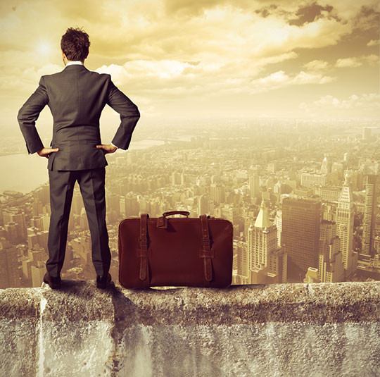 Взрослый мужчина в пальто стоит с сумкой на плече и чемоданом.