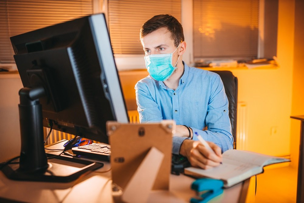 Взрослый мужчина сидит за столом с монитором на фоне мебели в офисе.