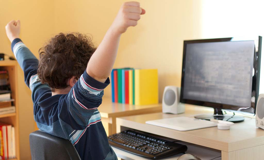 Мальчик-победитель радостно улыбается, сидя за монитором с аудиосистемой и клавиатурой.